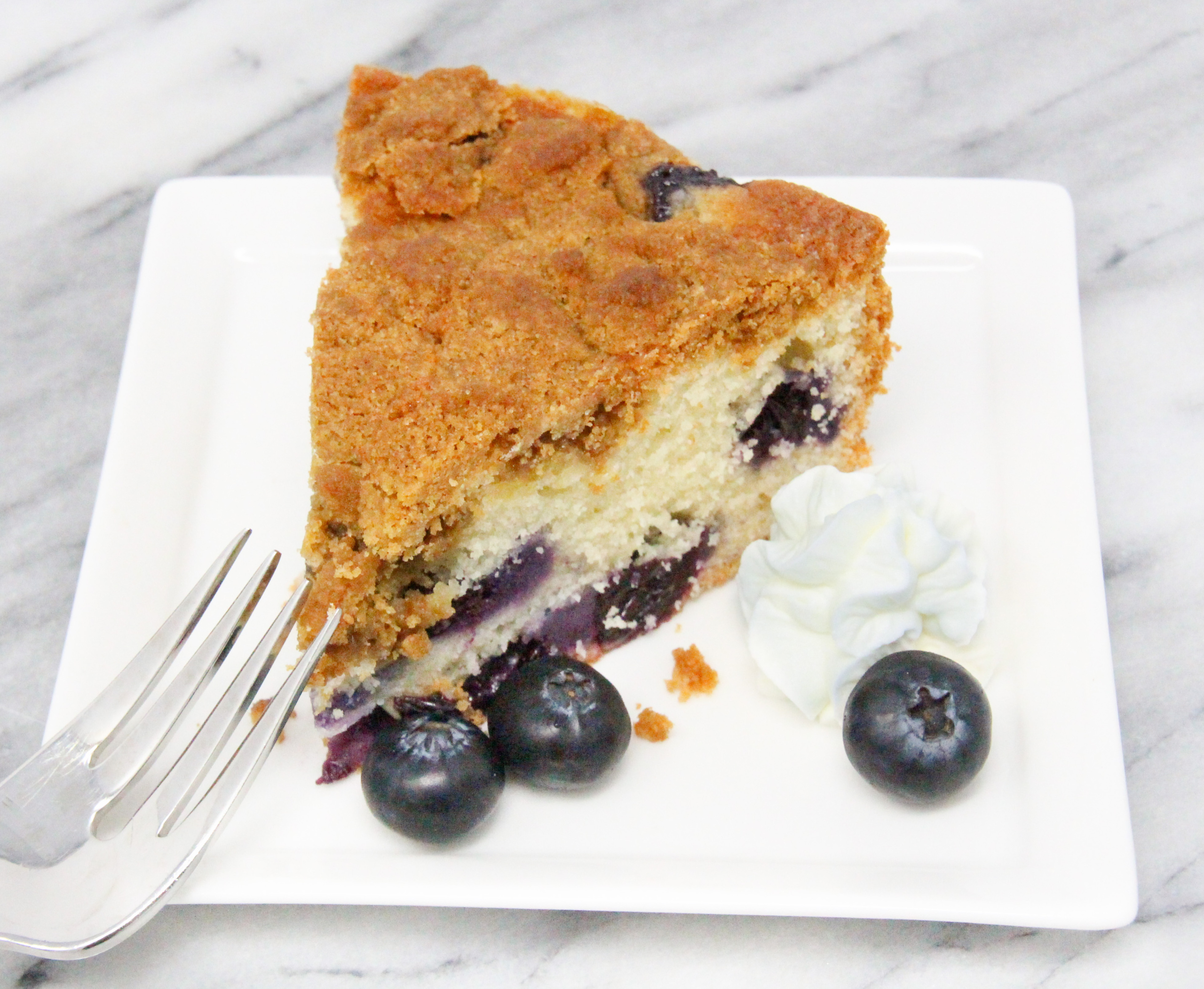 Blueberry Buckle has thick, crunchy streusel topping which enhances the sweet vanilla cake studded with fresh blueberries. With a dollop of freshly whipped cream, this really is darn fine! Recipe shared with permission granted by Amy Pershing, author of MURDER IS NO PICNIC.