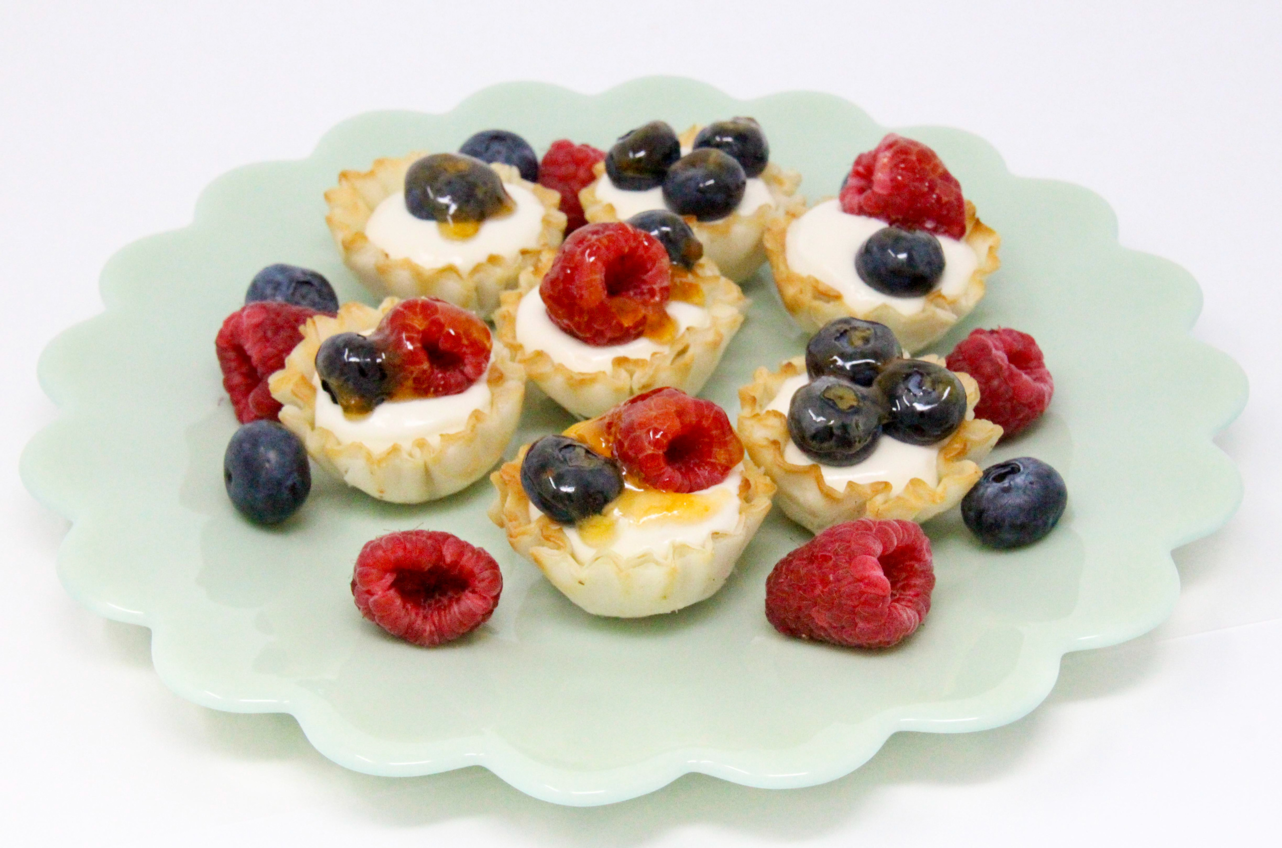 Mini No-Bake Honey Cheesecakes starts with the ease of premade mini phyllo cup shells and filled with honey sweetened no-bake cheesecake filling. Topped with berries, these make for a light and satisfying treat. Recipe shared with permission granted by Kim Davis, author of ESSENTIALS OF MURDER. 